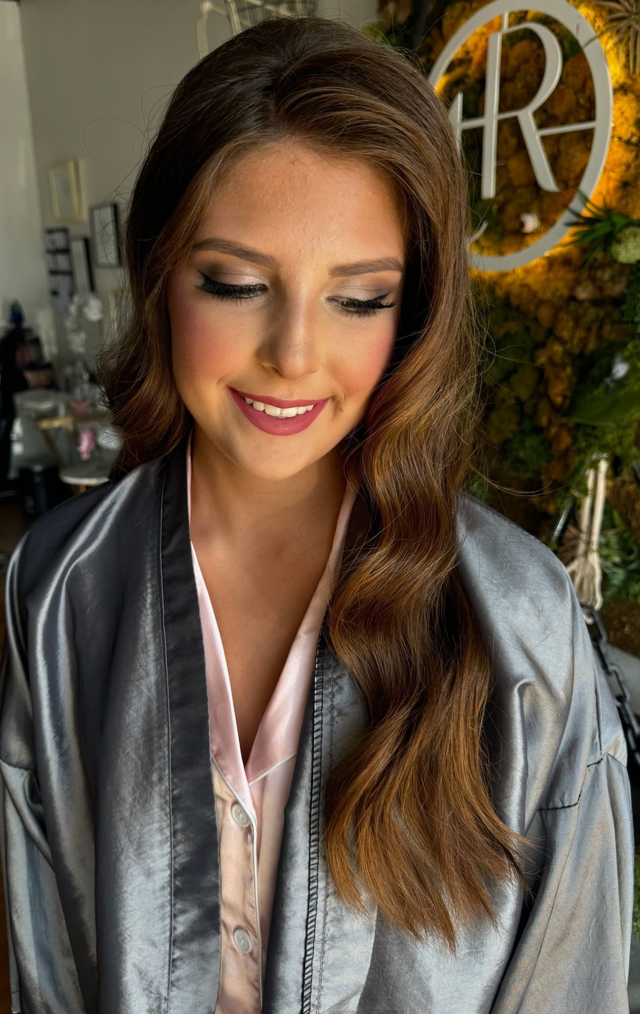 Woman with brown hair and makeup, wearing a grey robe and pink shirt, looking down.