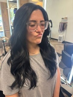 Woman with dark wavy hair wearing glasses puckers her lips in a salon.