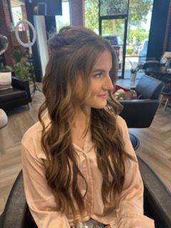 Woman with wavy brown hair in a salon, wearing a pink shirt, smiling.