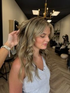 Woman with wavy, light blonde hair styled at a salon. Someone's hand is in her hair.