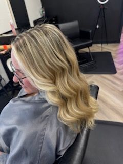 Woman with wavy blonde hair, highlights. Sitting in a salon chair, wearing glasses and a gray smock.