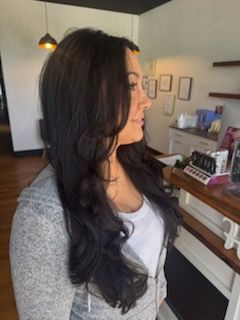 Woman with long, dark wavy hair in a salon wearing a grey sweater and white shirt.