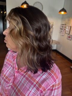 Woman with wavy hair, a mix of dark and blonde, in a pink plaid shirt, facing left in a room with wall decor.