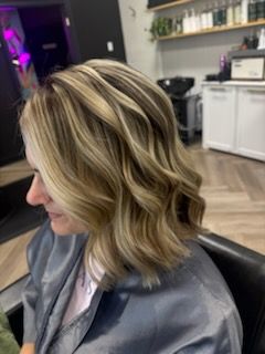 Woman with wavy blonde hair highlights in a salon setting, wearing a gray robe.