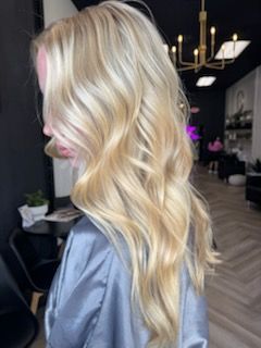 Woman with long, wavy blonde hair. Wearing a grey satin robe in a salon.