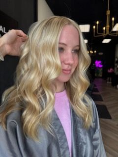 Blonde-haired woman with wavy hair, wearing a pink shirt and gray robe, inside a salon.