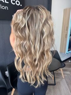 Woman with long, wavy blonde hair.  Facing away, wearing black clothing, in a salon setting.