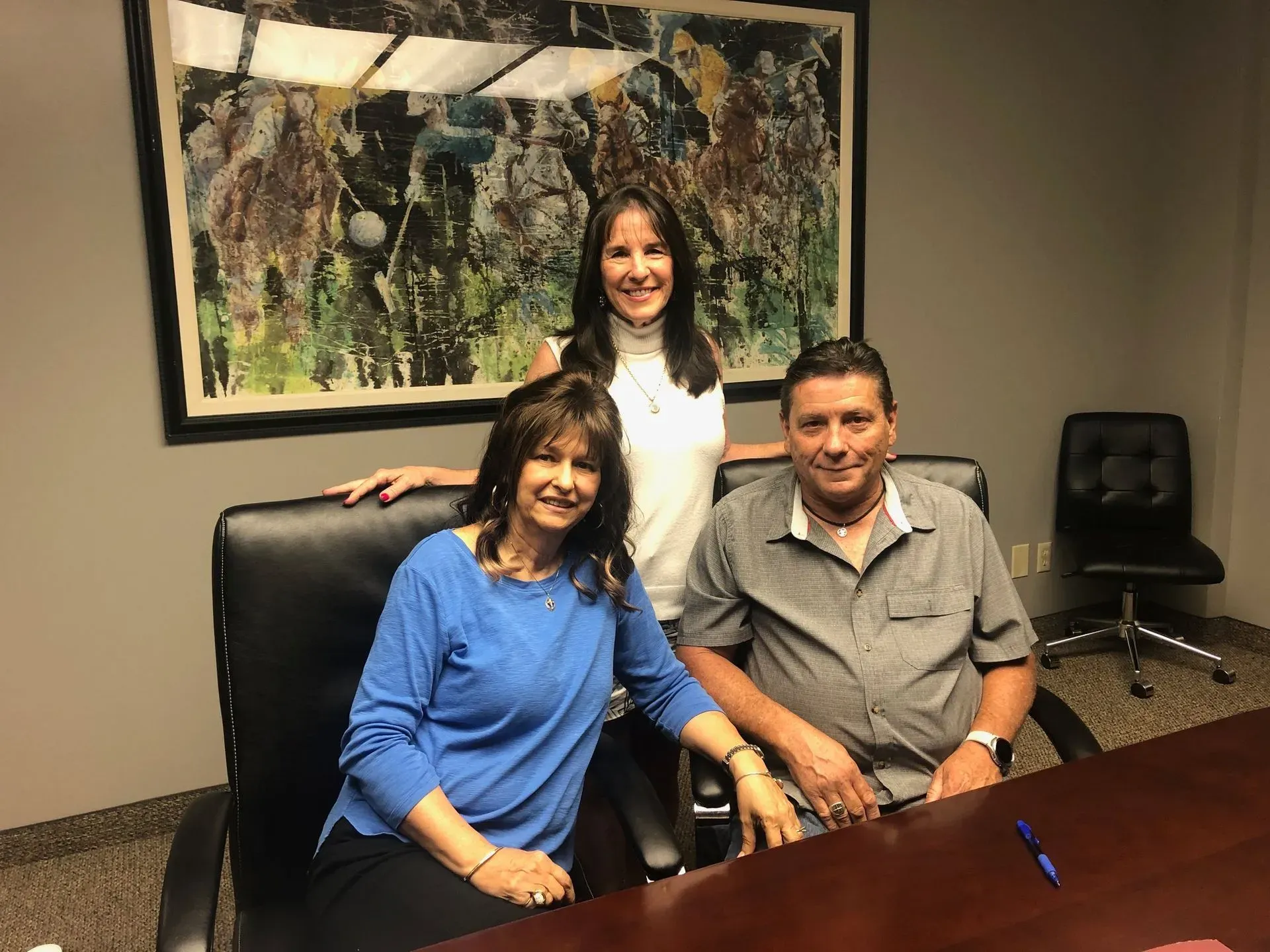 Two smiling individuals sit at a conference table while a third stands behind them in front of an abstract framed artwork.