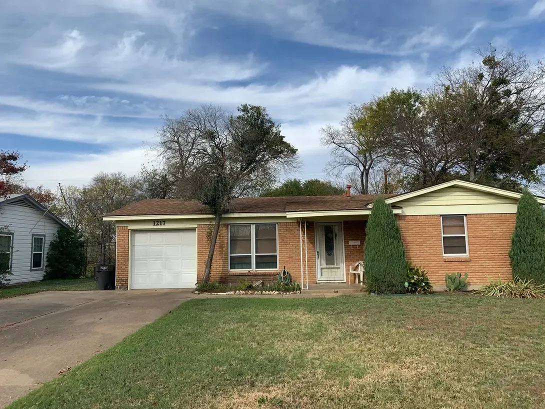 A single-story brick house with a white garage door, light siding, and two tall evergreen bushes in a sunny yard.