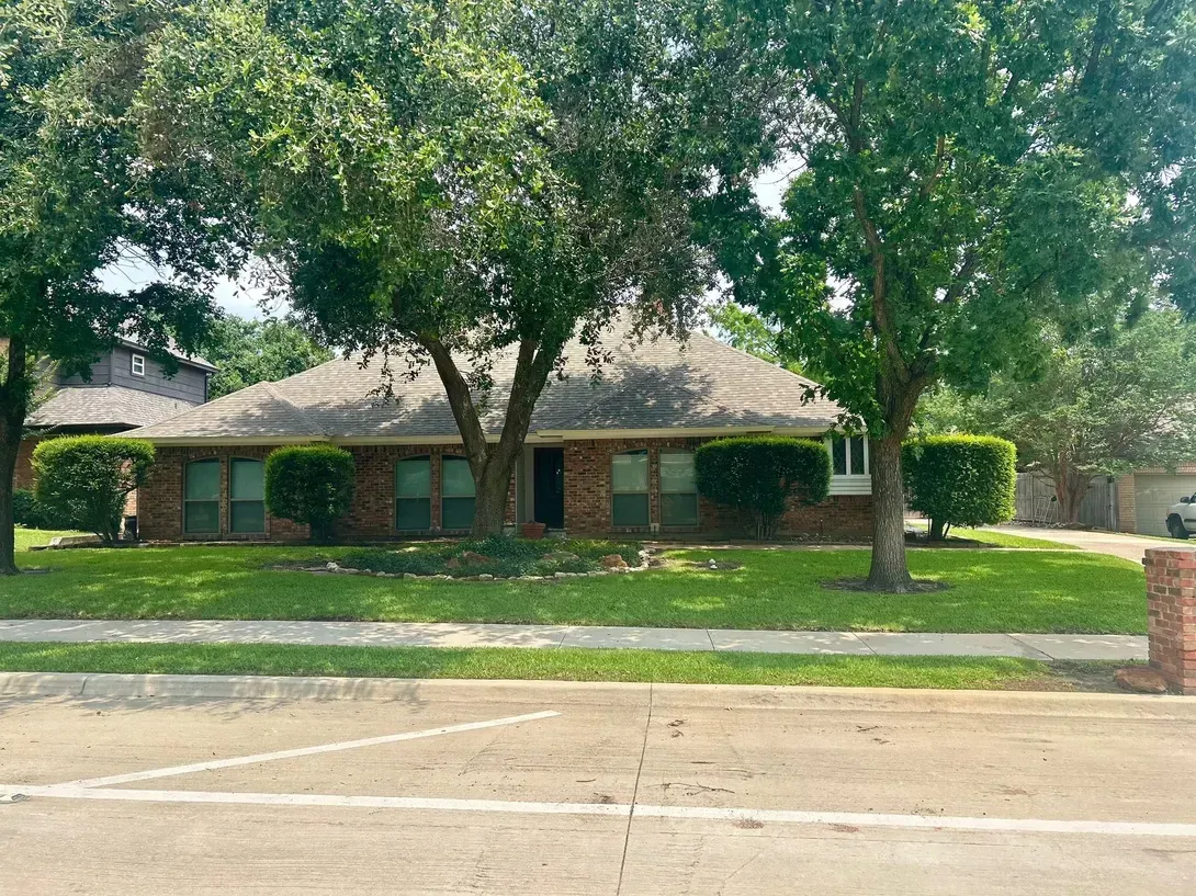 A single-story brick house with a gray shingled roof, surrounded by green lawns and mature shade trees.