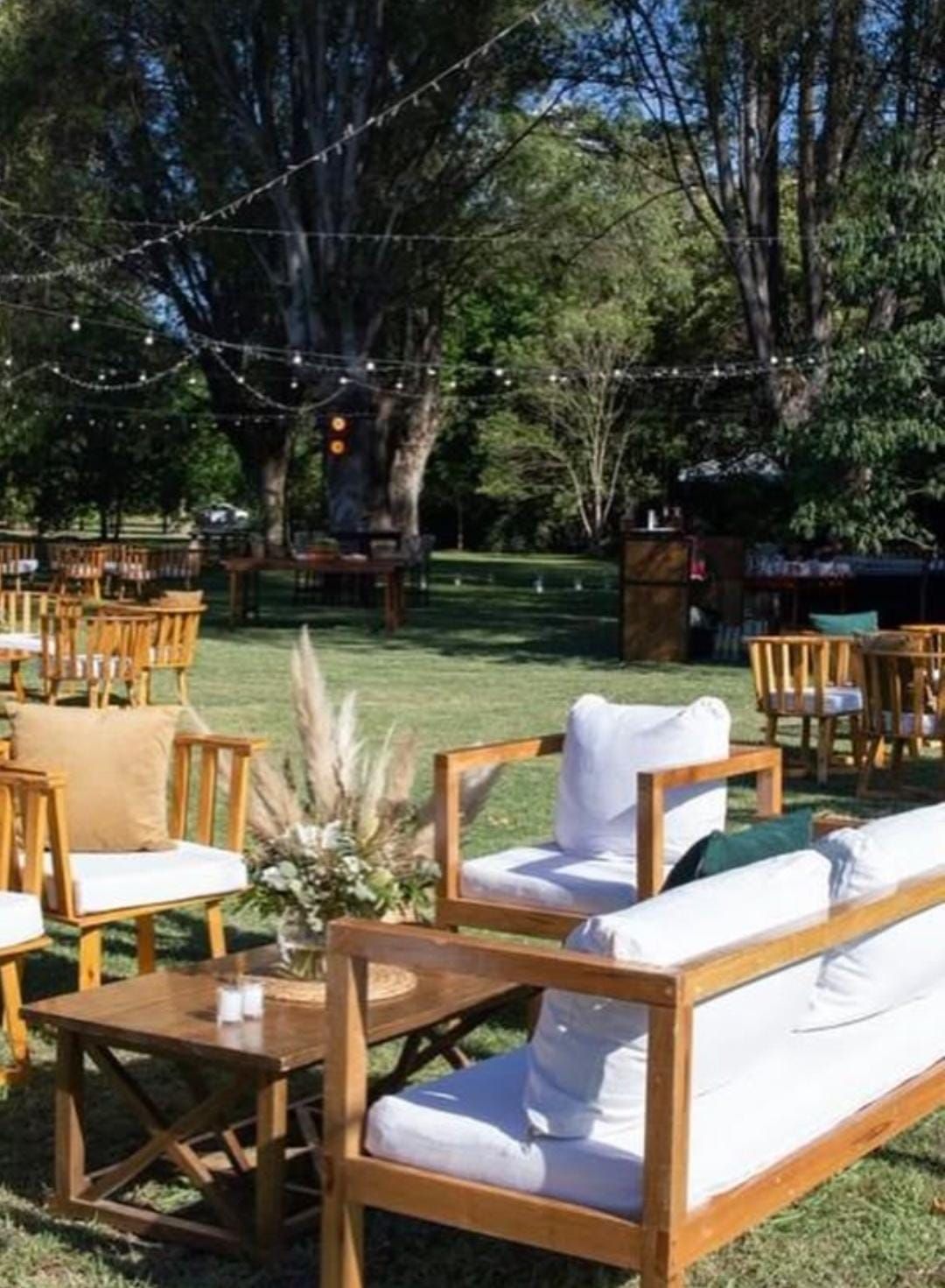 Zona de estar al aire libre con muebles de madera, cojines blancos y luces de cadena, ubicada en un patio con césped.