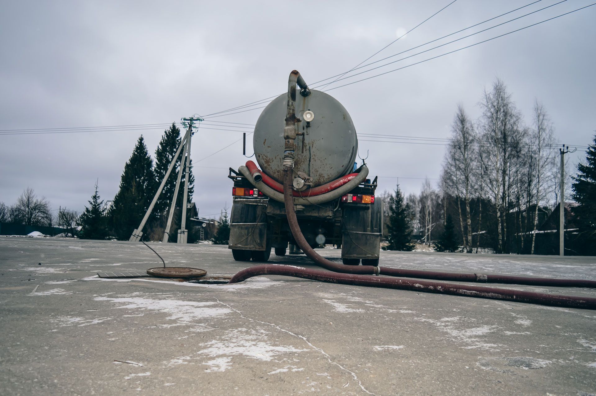 Tanker truck pumping waste from a manhole on a concrete surface. Gray sky, leafless trees.