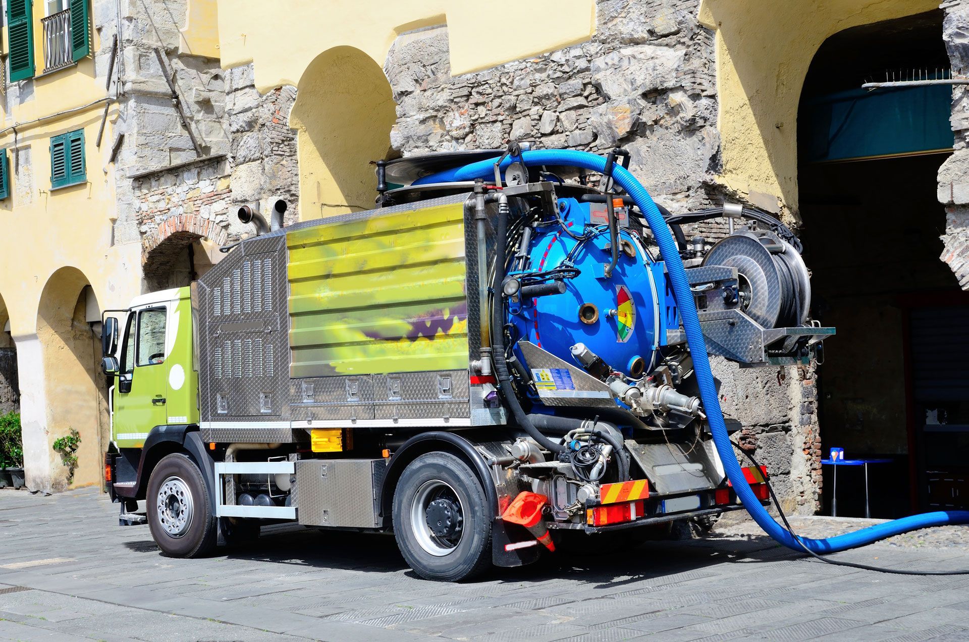 Green and silver sanitation truck parked on cobblestone street. Blue hose and tank visible.
