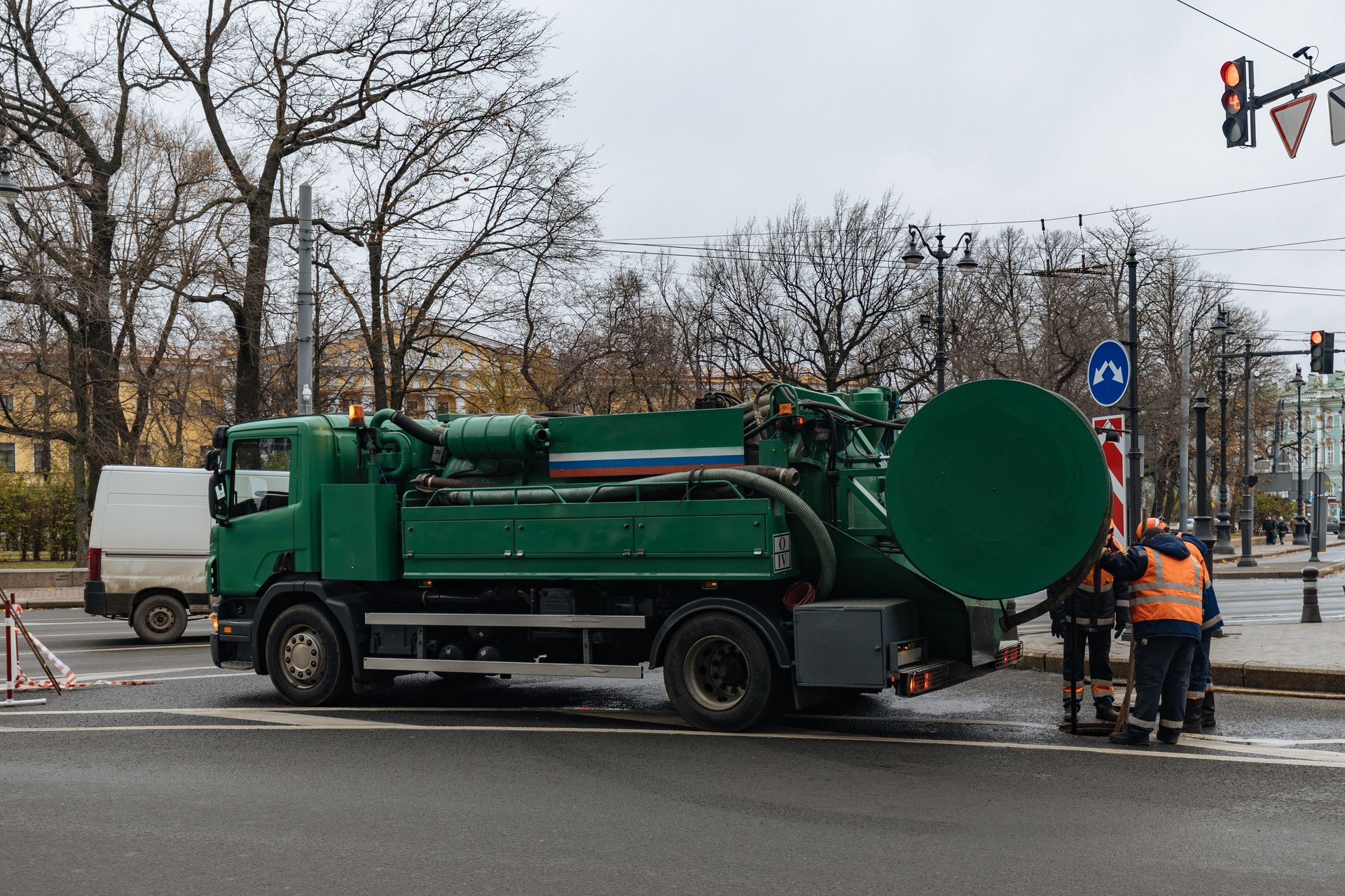 Green sewage truck on road; workers near sewer entrance. Trees and buildings in background.