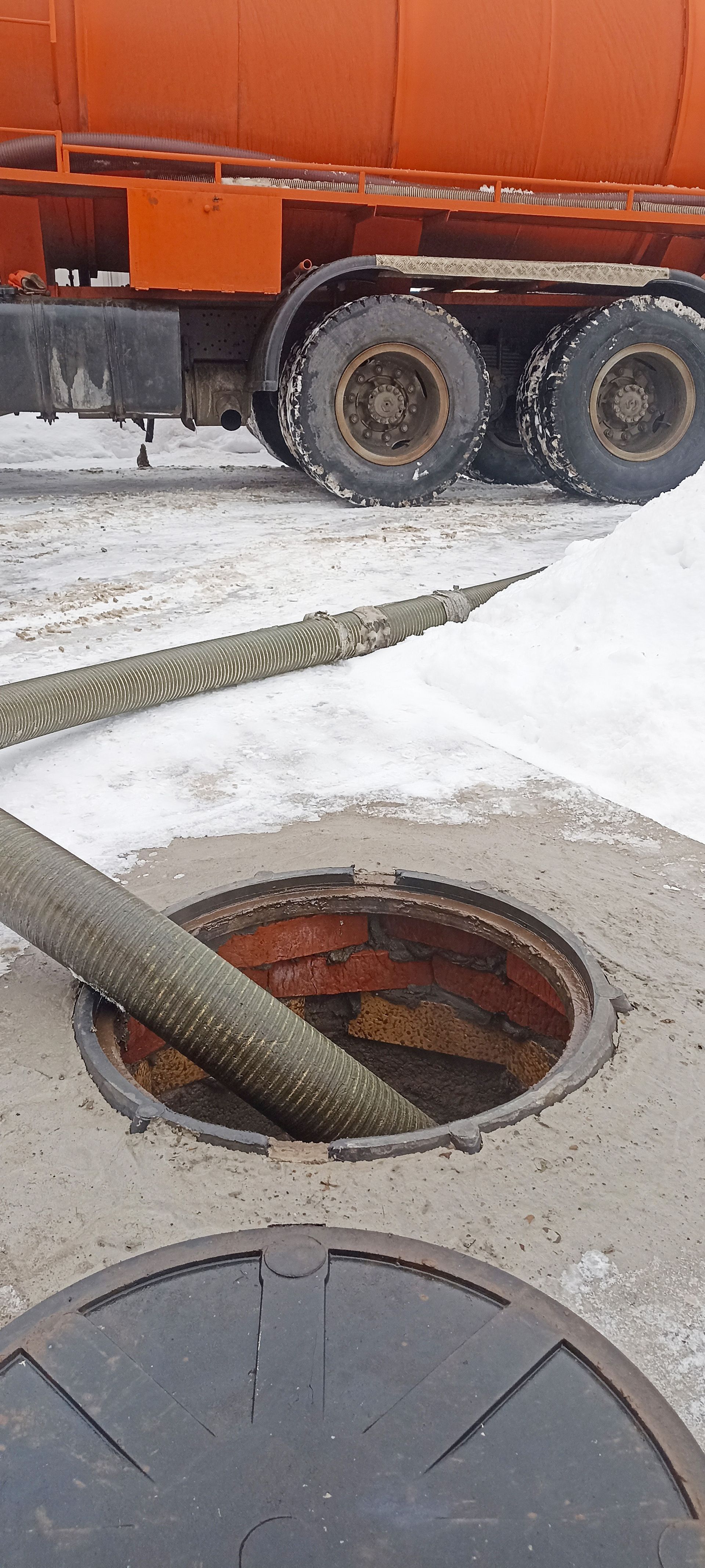 An orange tanker truck with a hose connected to a manhole in the snow.