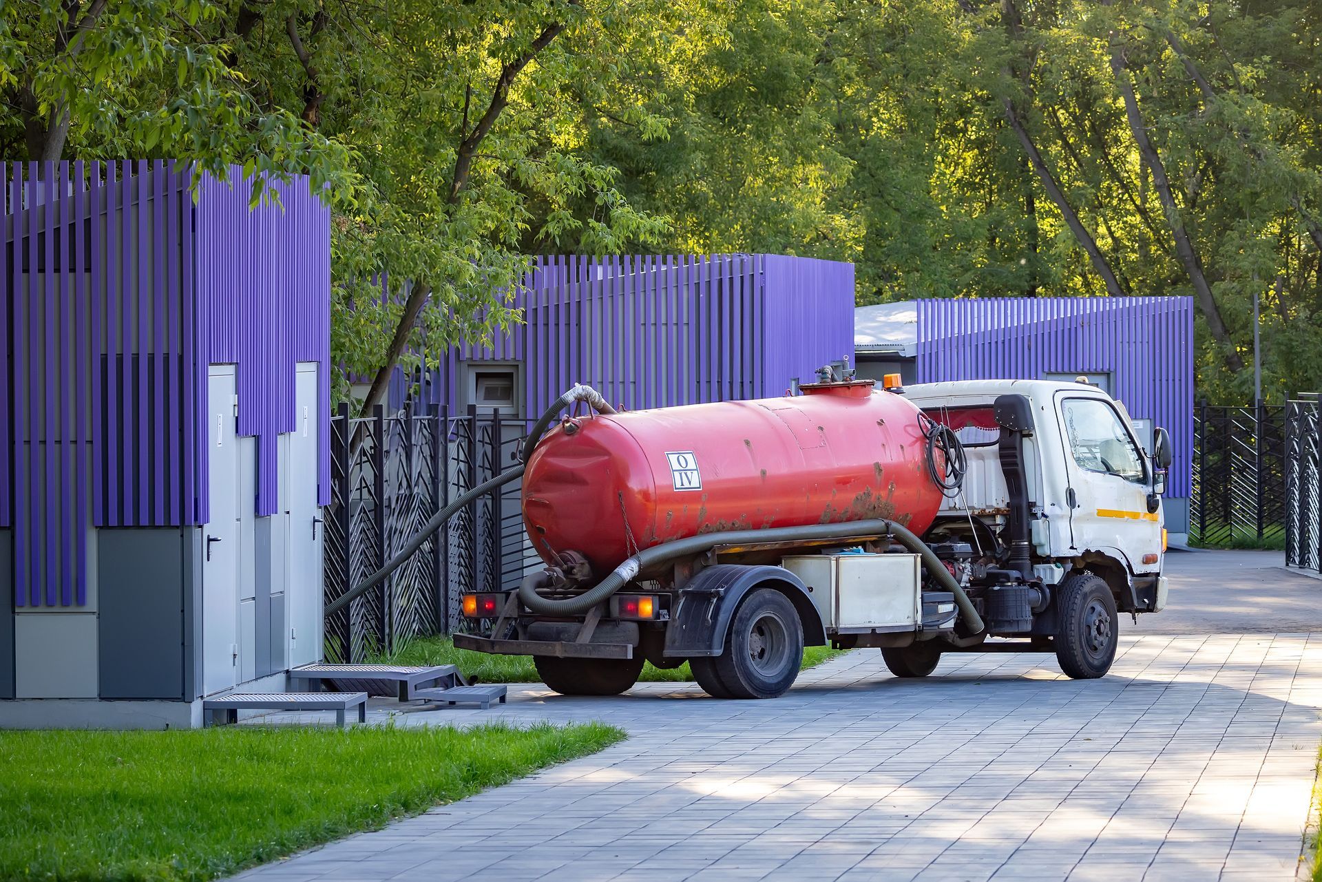 Red septic tank truck parked next to purple fence, taking waste. Outdoors, trees in background.