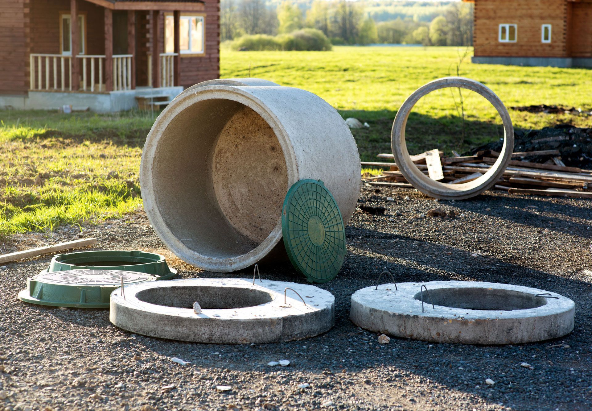 Concrete well components outdoors: rings, cylinder, and covers, near buildings and green grass.