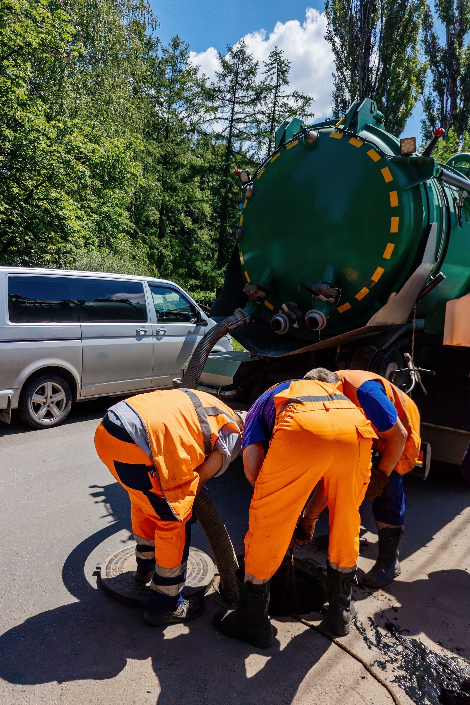 Three workers in orange vests connecting hose to sewer drain, with a large green tank truck nearby.