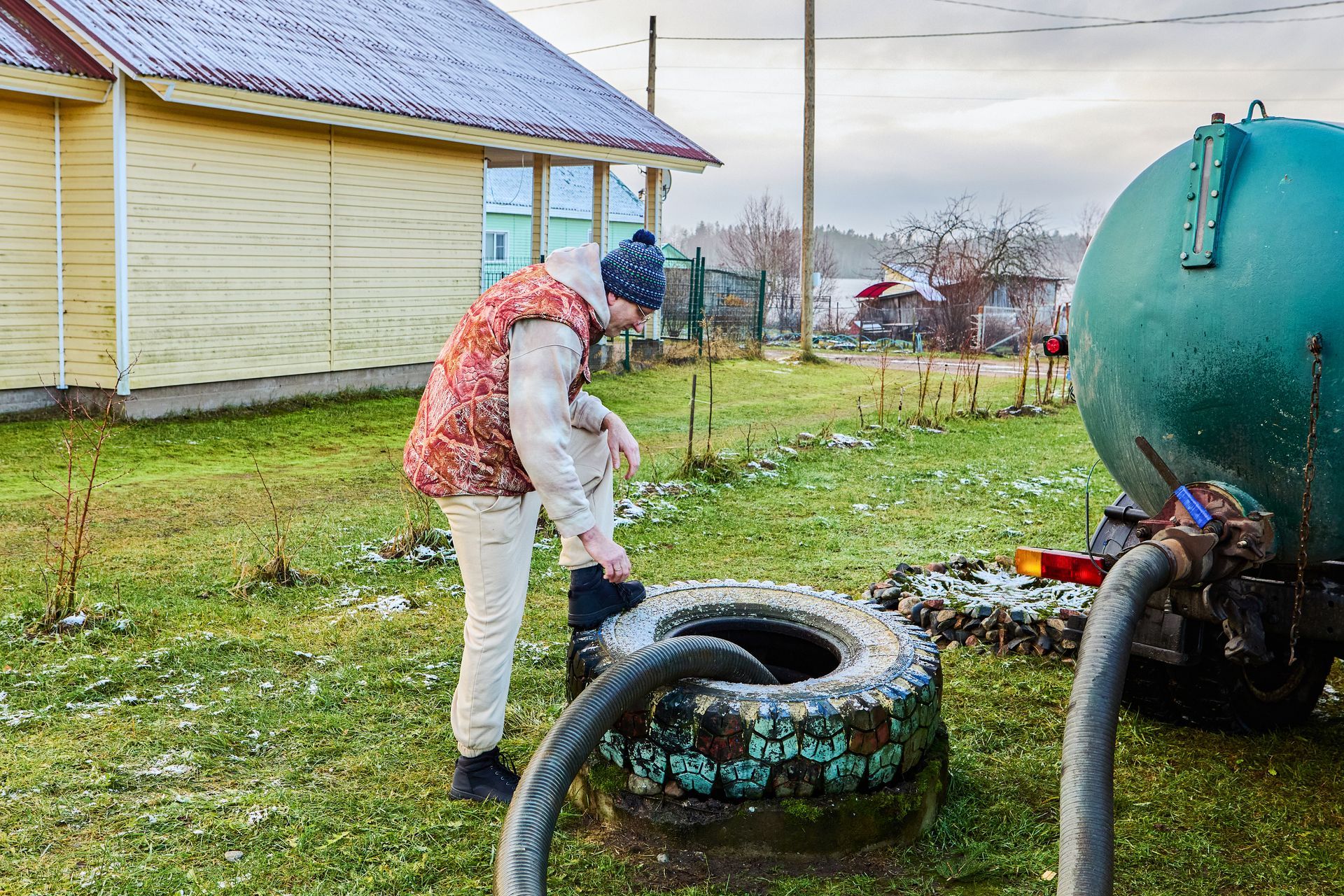 Person in a red vest and white apron tending a large hose by green tanks in a muddy yard.
