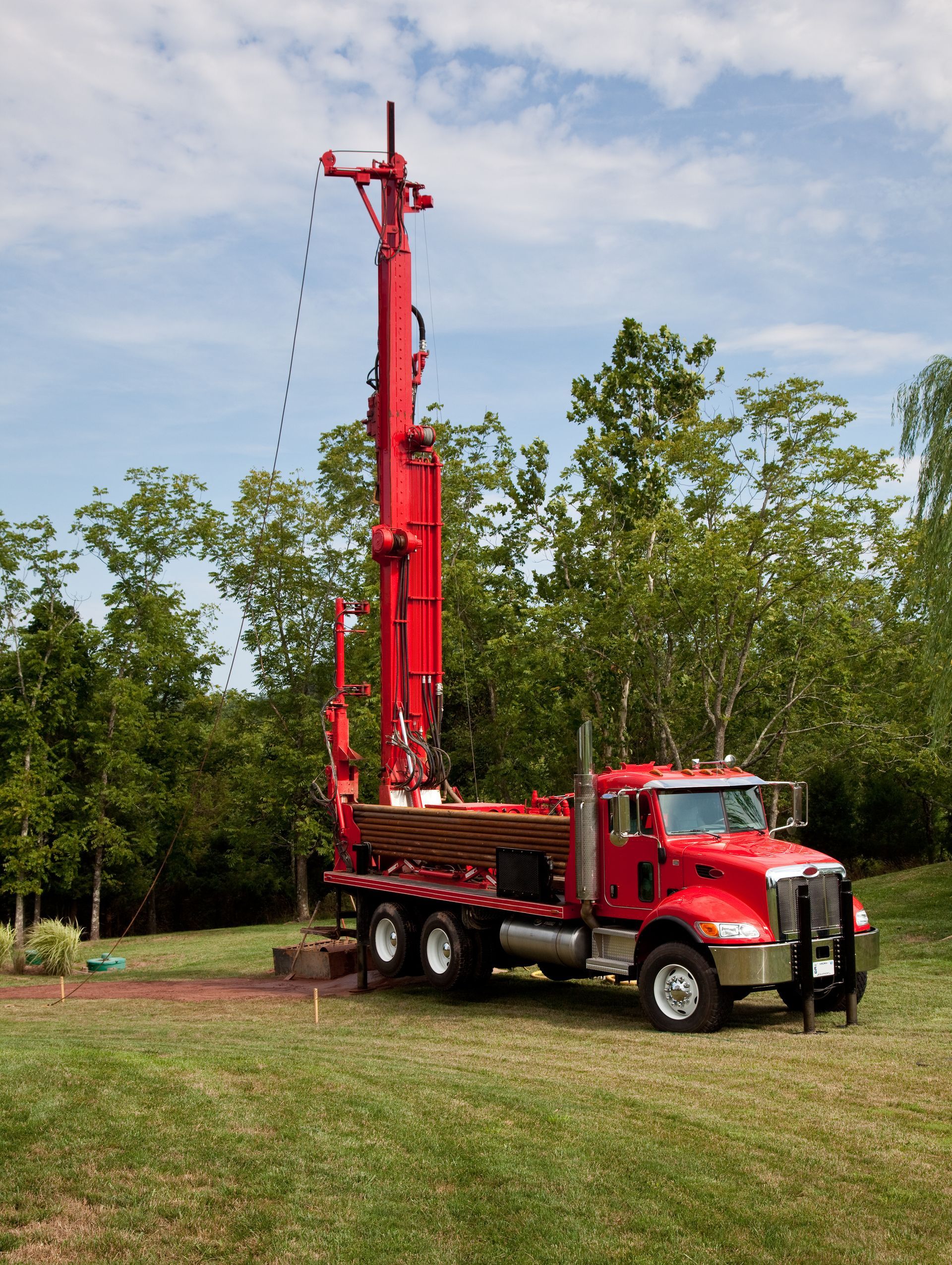 Red drilling rig on a truck in a grassy area with trees, under a cloudy sky.