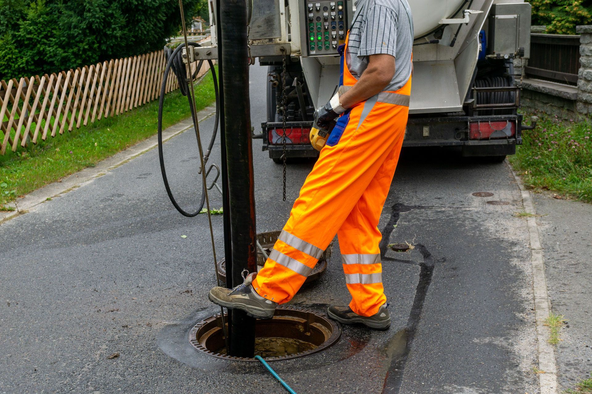 Worker in orange overalls standing over an open manhole on a street, with a truck in the background.