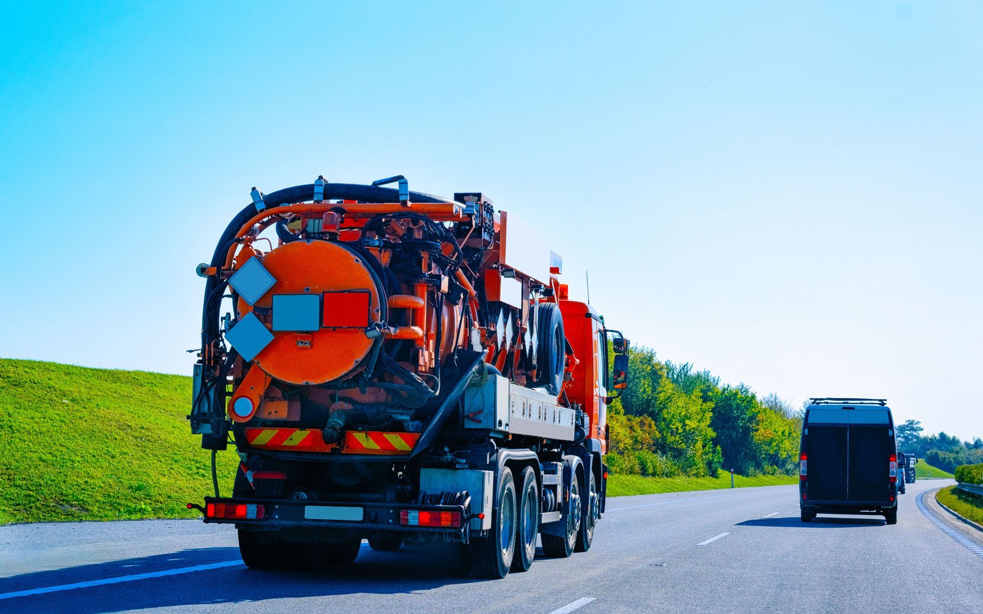 Rear view of an orange and black vacuum truck driving on a highway, with a smaller vehicle in the background.
