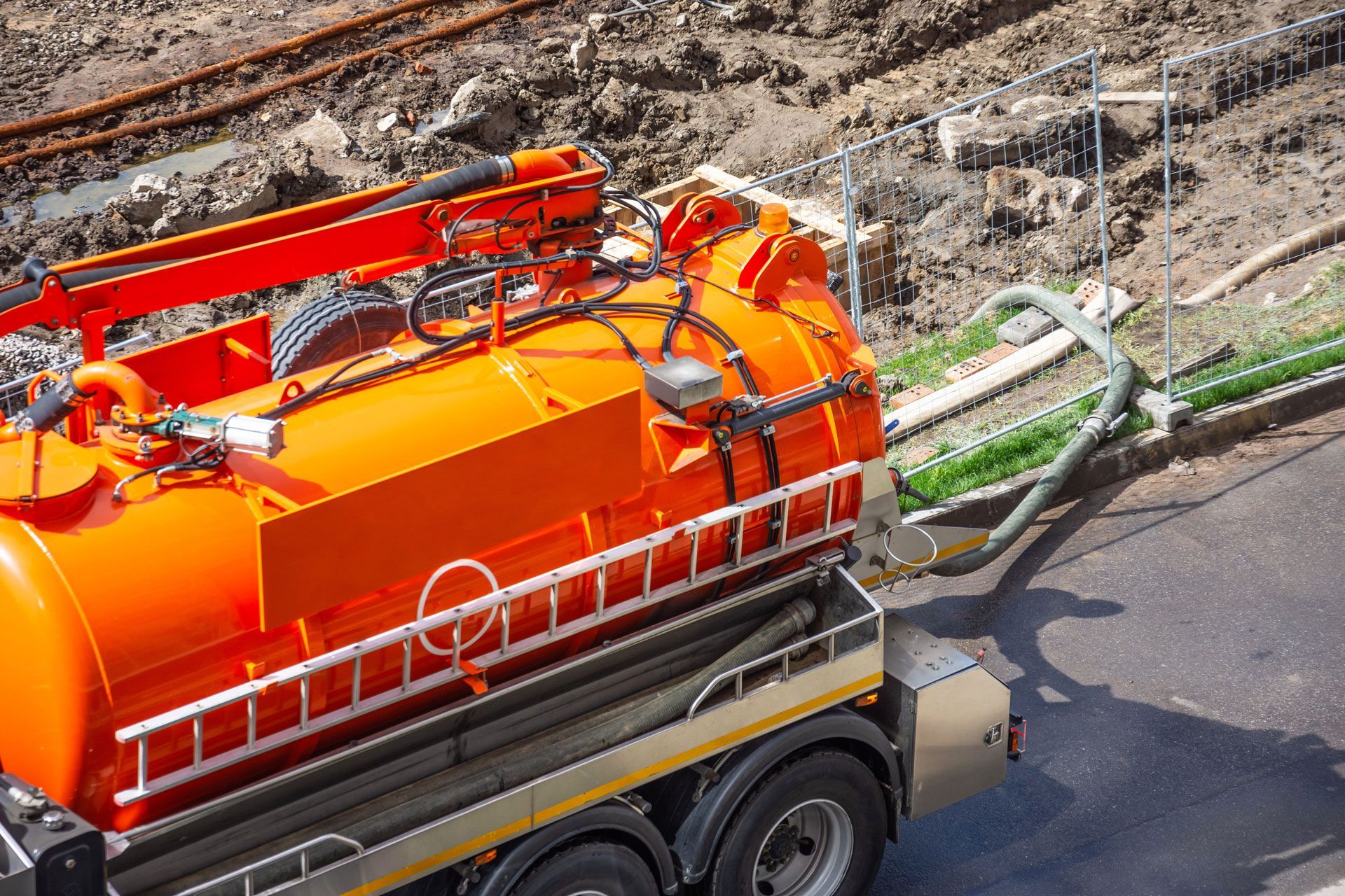Orange vacuum truck, hoses connected, parked near construction site with dirt and pipes.