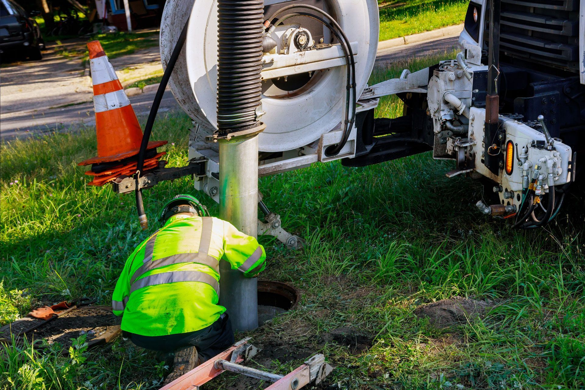 Worker in neon vest near a utility hole, operating equipment from a truck with a cone on grass.