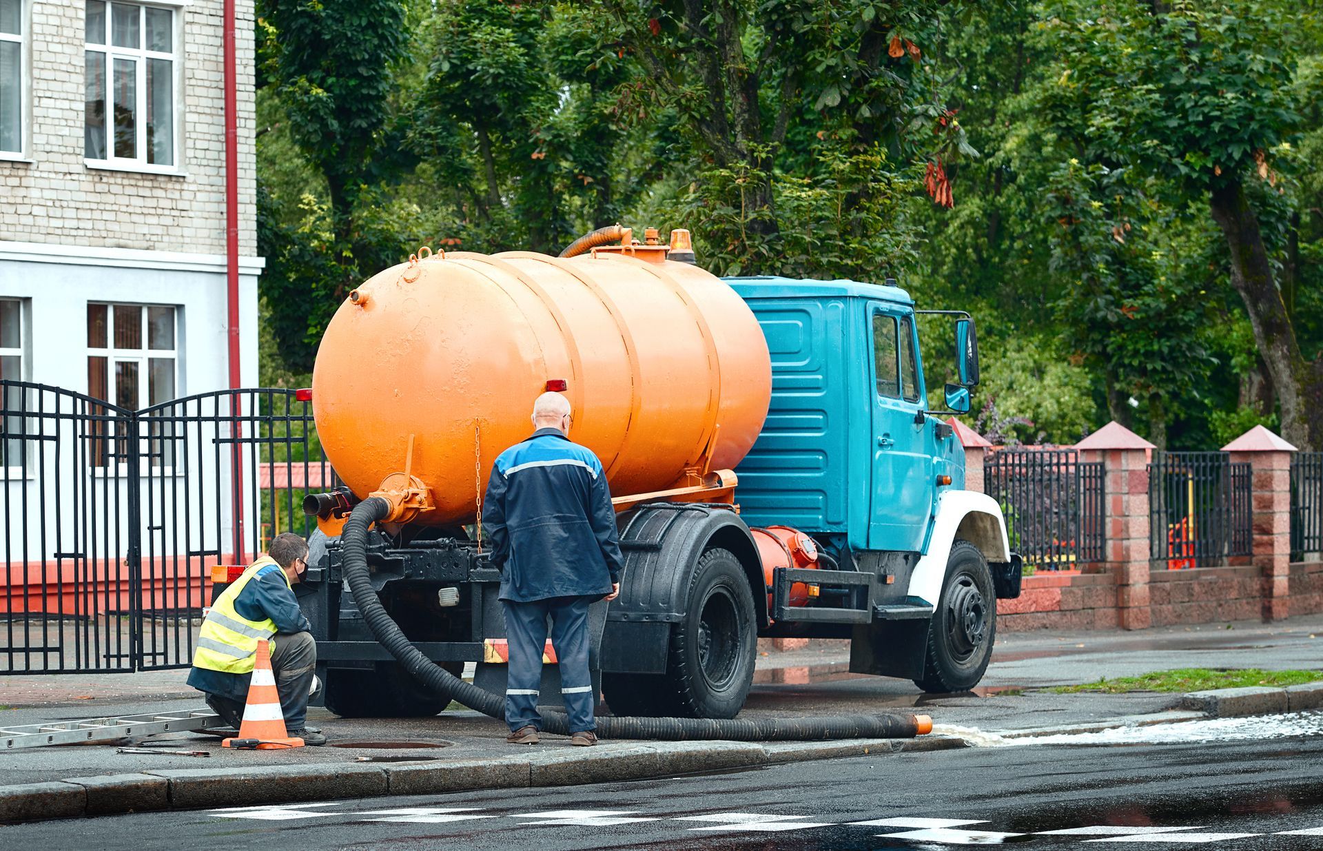 Workers with a blue and orange tank truck are working on a street next to a building.