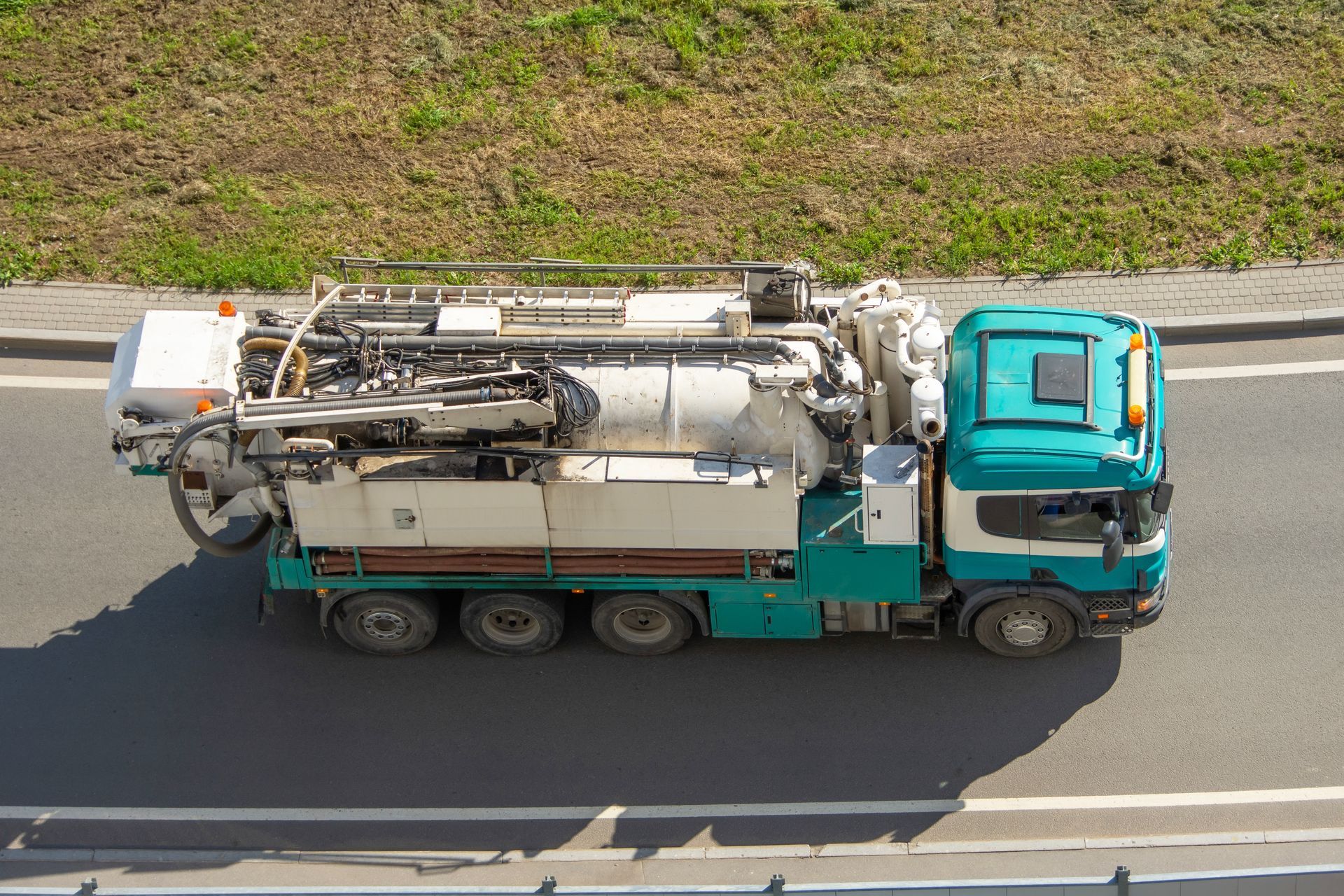 Green and white vacuum truck on a road, operating near a grassy slope.