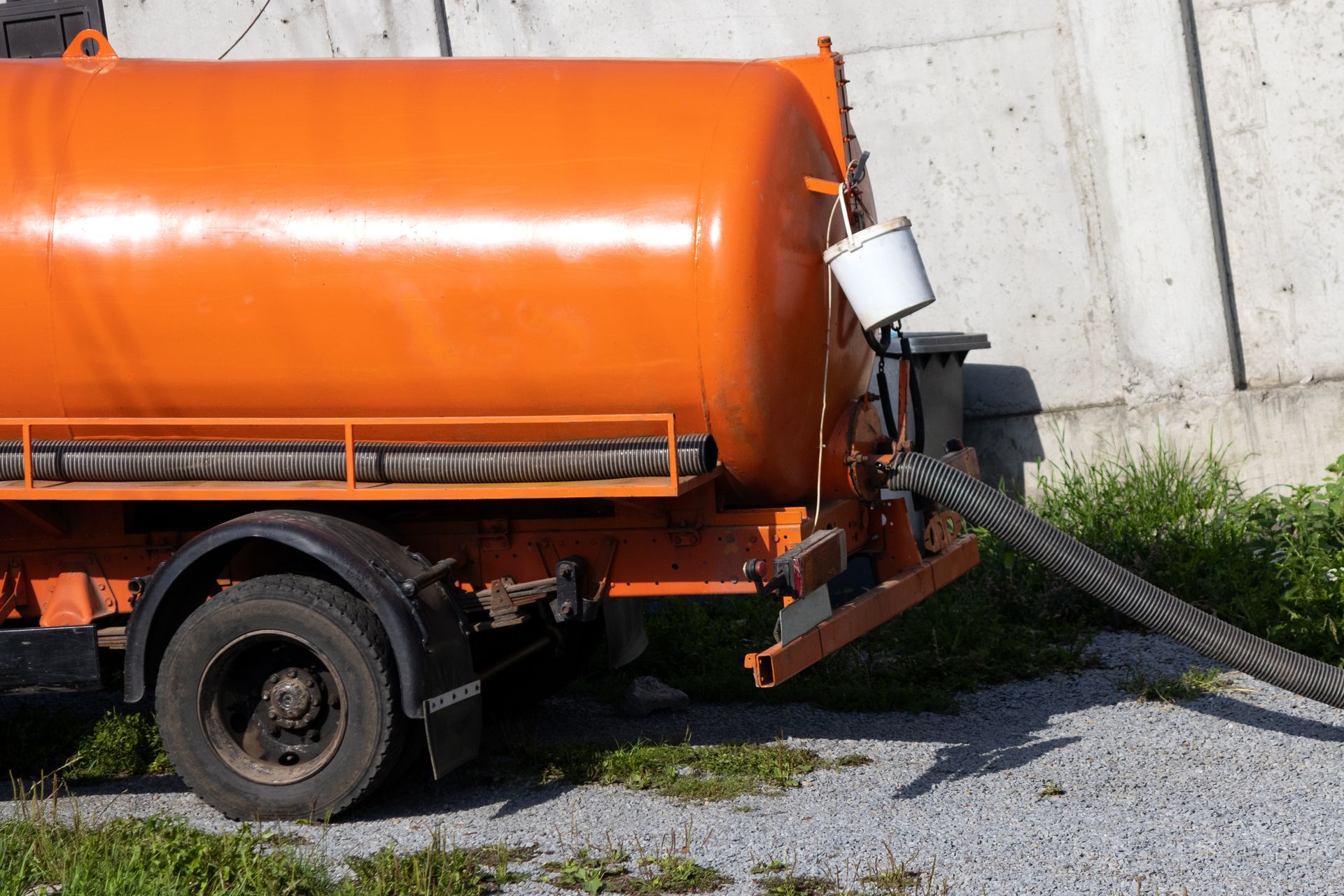 Orange tanker truck parked near a concrete wall, hose extended onto gravel.