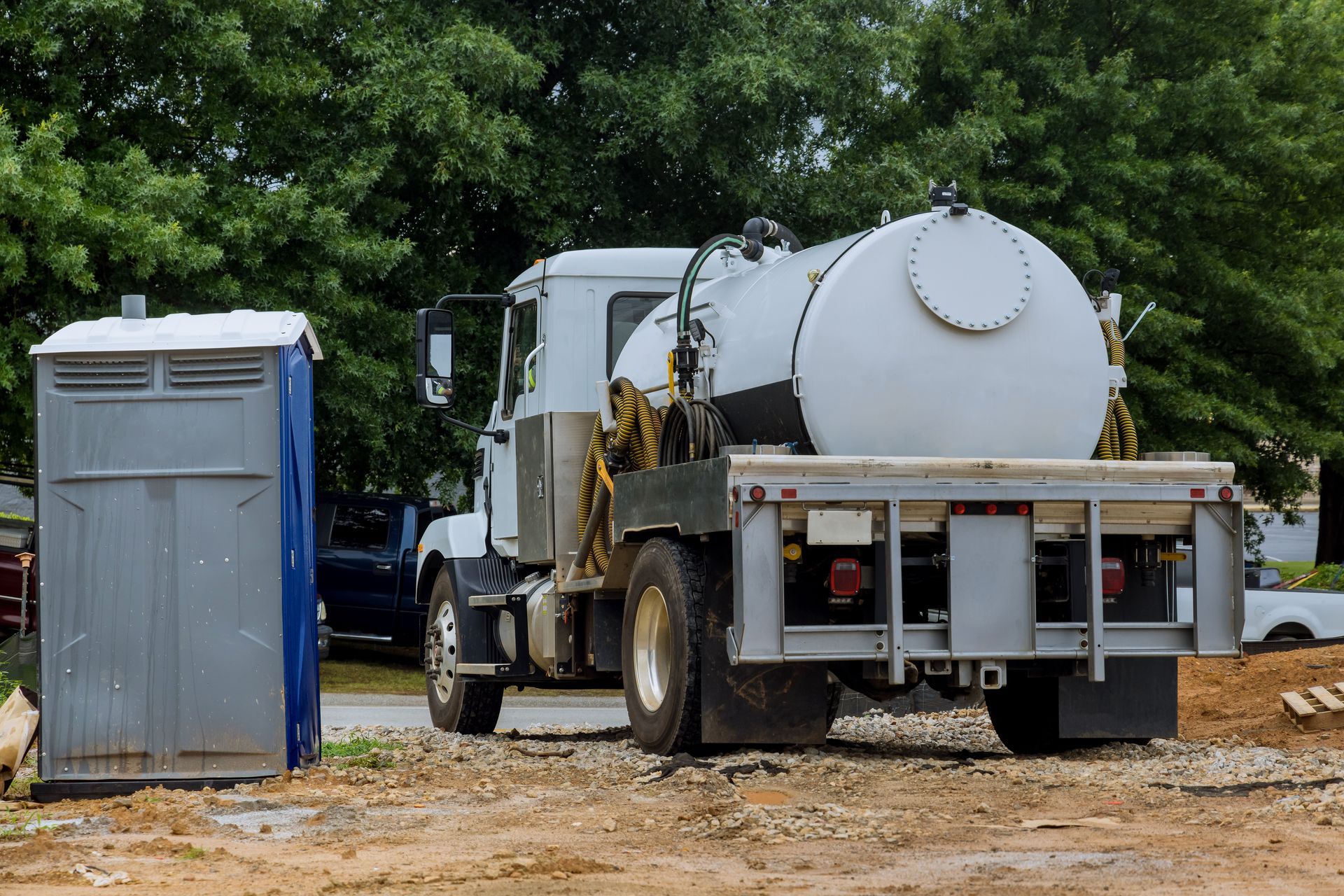 Portable toilet next to a truck with a large tank, parked on dirt.