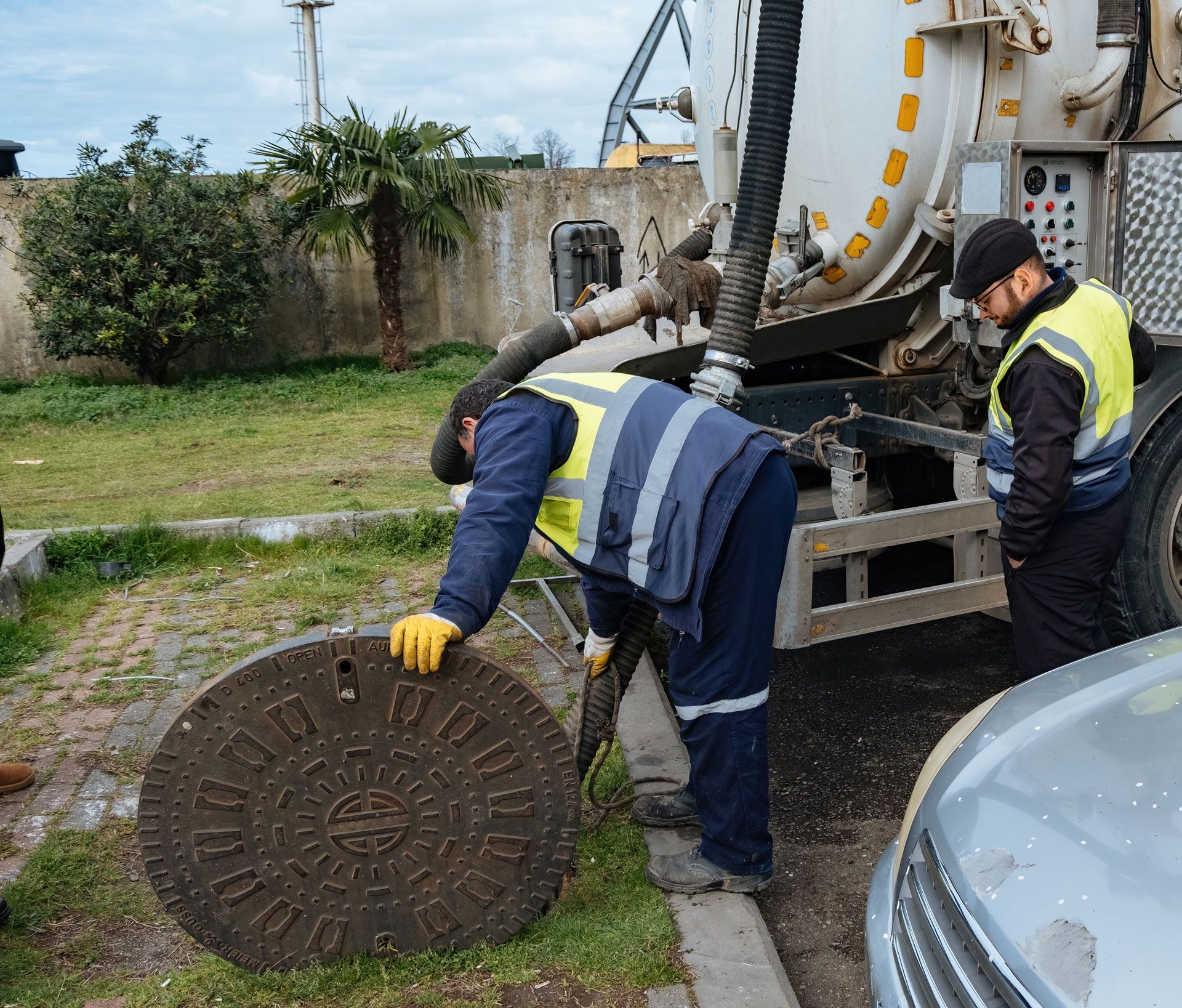 Two workers near a truck. One lifts a manhole cover. The other observes.