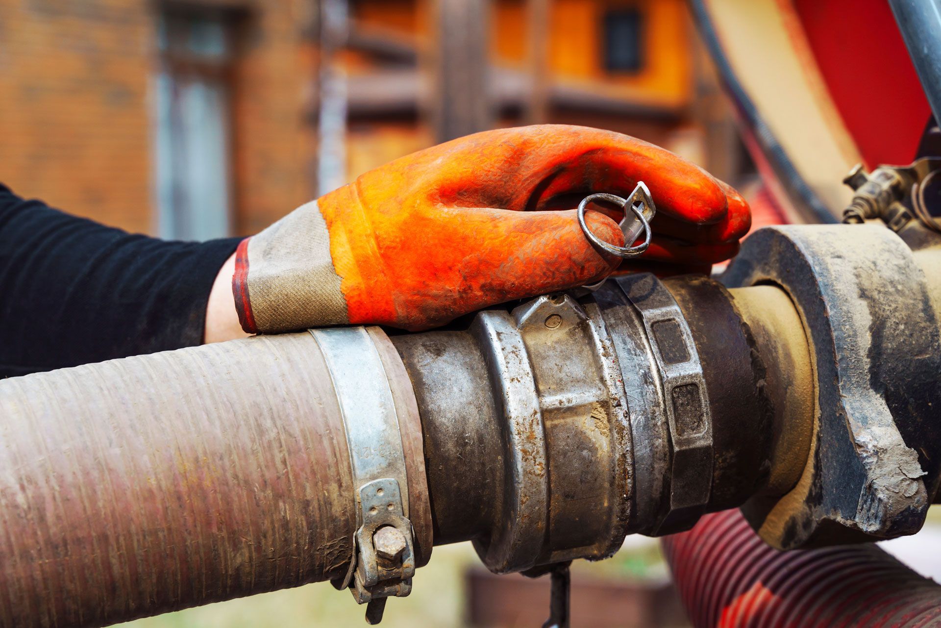 Gloved hand rests on a dirty pipe connection with clamps. Orange glove, silver ring.