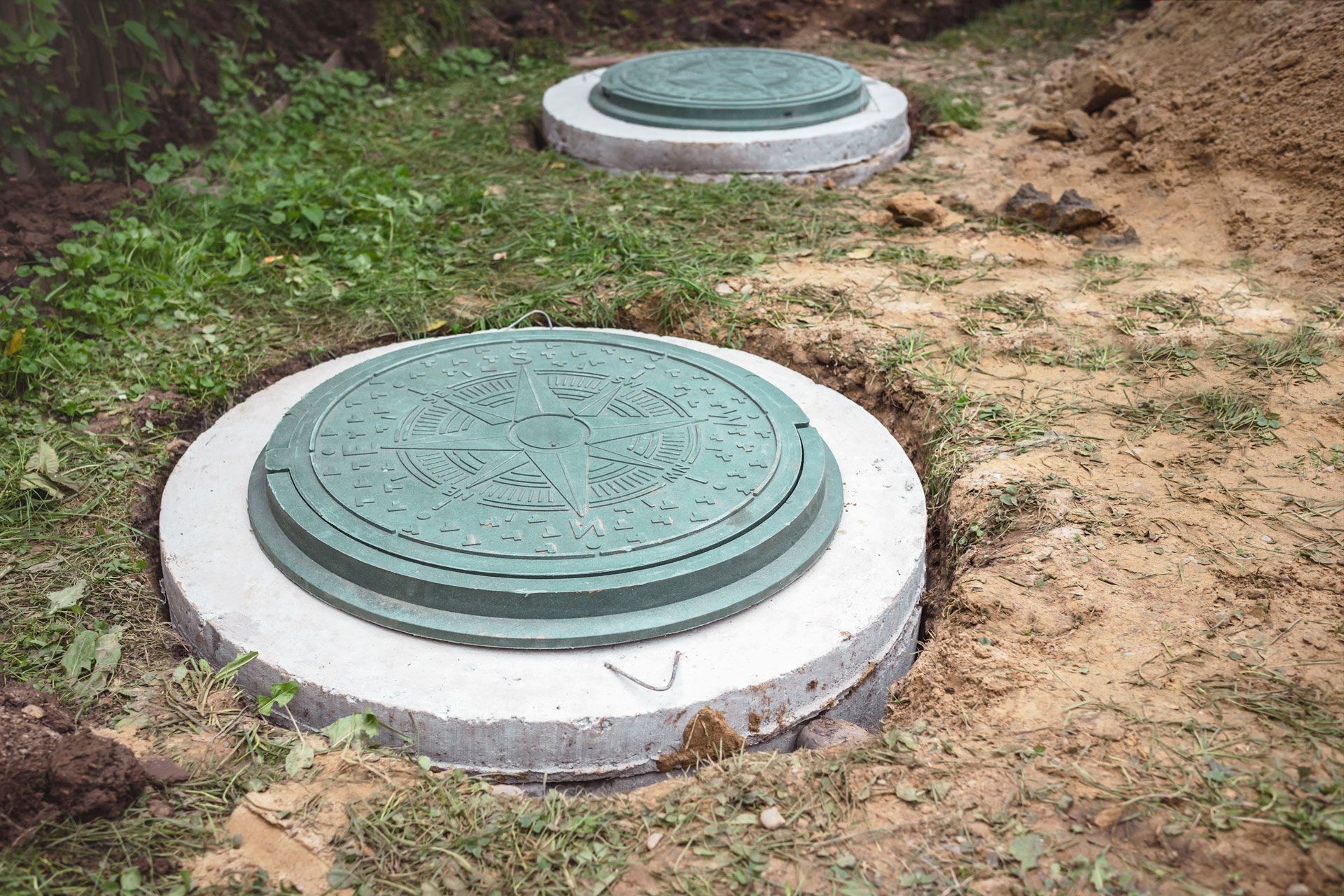 Two green septic tank lids set in concrete rings, surrounded by dirt and grass.