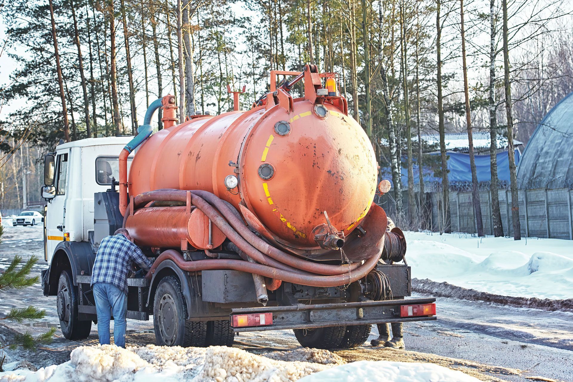 Orange septic tank truck with a person working on it, parked in a snowy area.