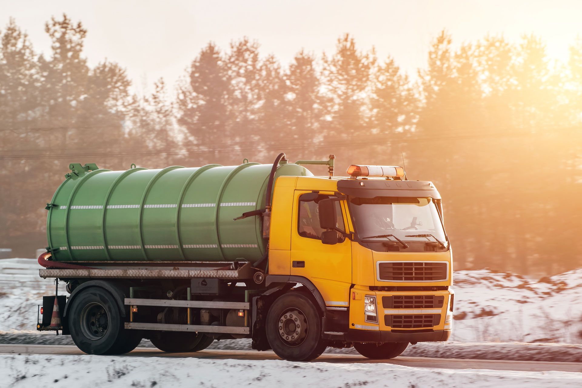 Yellow septic truck with green tank on a snowy road, trees in the background, sunlit scene.