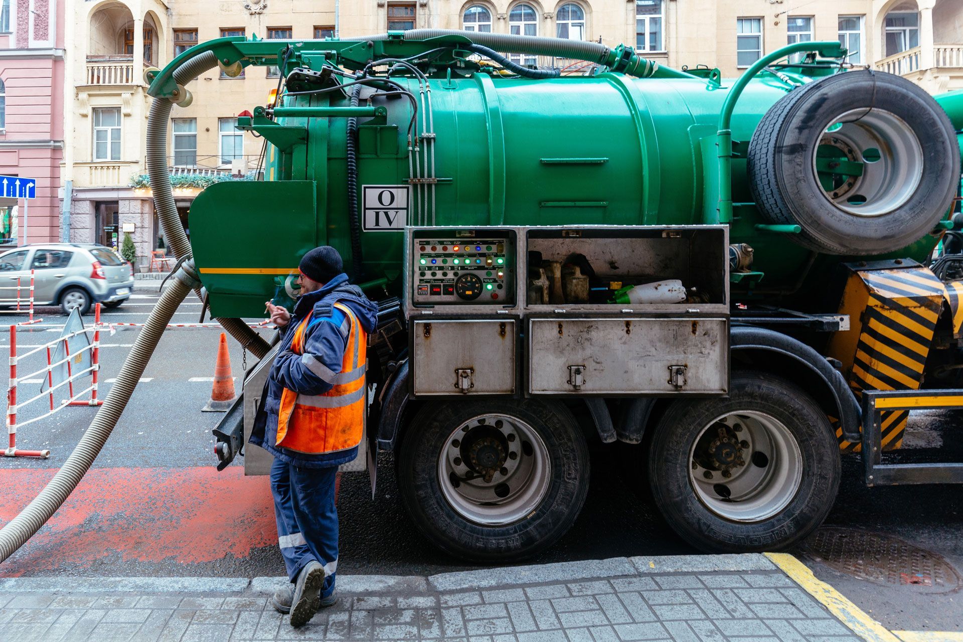 Worker in orange vest near green sewage truck on city street.