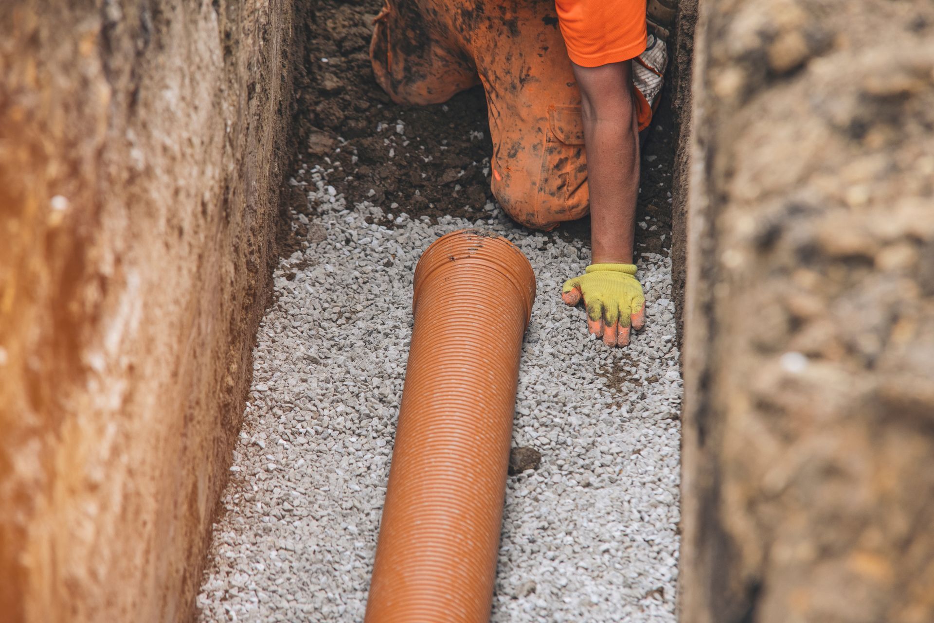 Worker installs orange pipe in a trench filled with gravel. Person kneels, wearing orange coveralls and gloves.