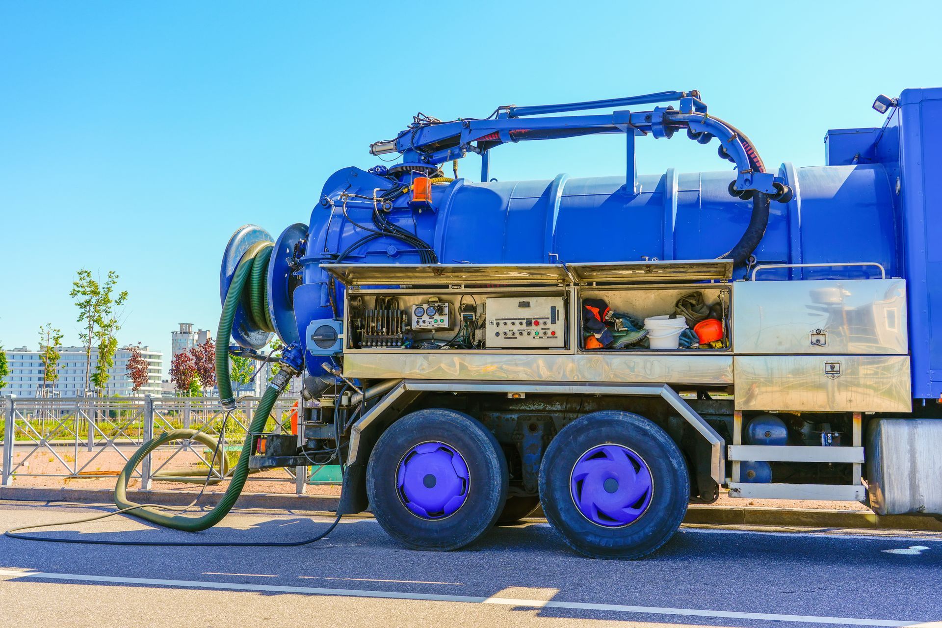 Blue vacuum truck on asphalt, bright sky. Equipment visible, hose extended.
