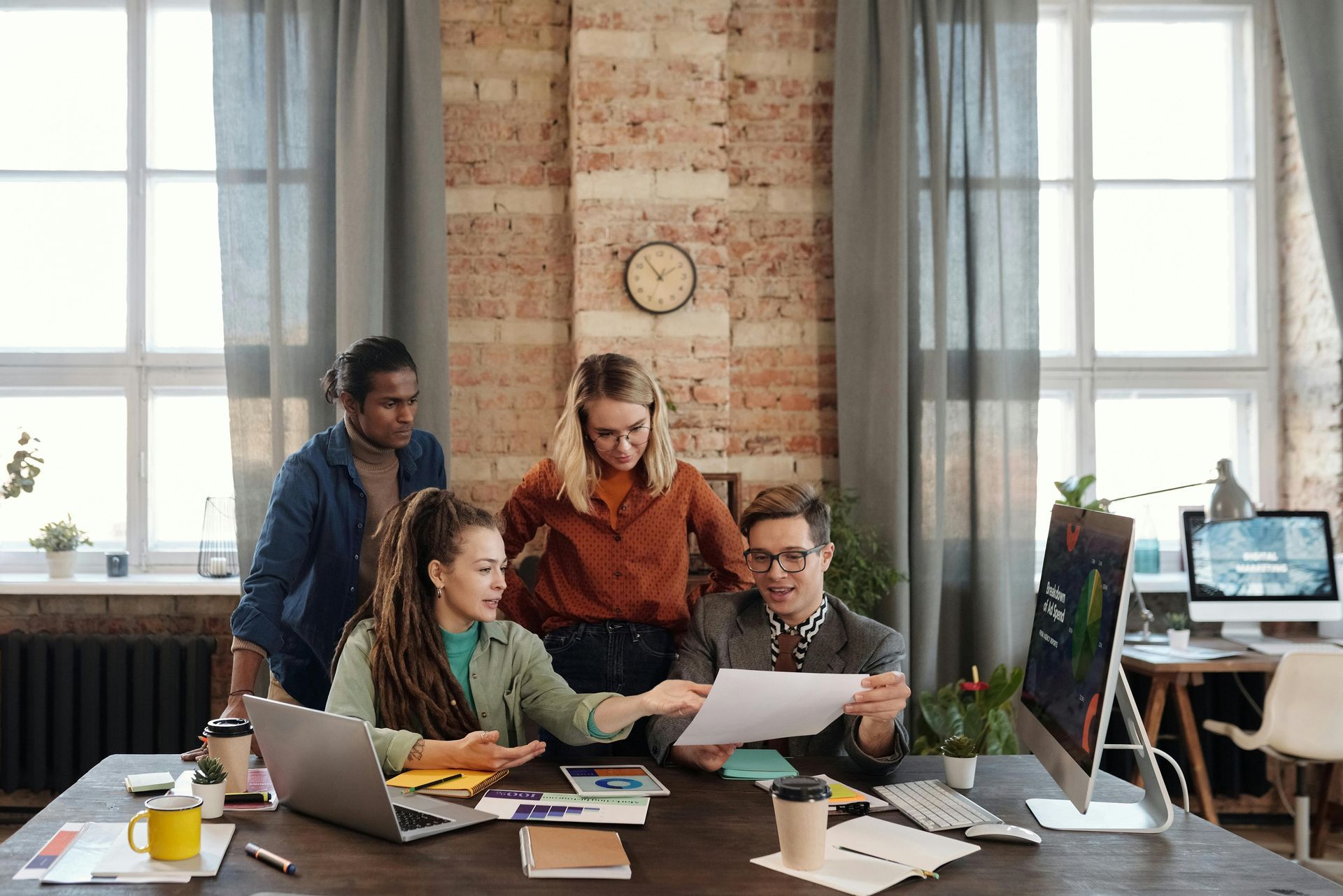 Four diverse colleagues collaborating around a table in a brick-walled office, reviewing papers and using a computer.
