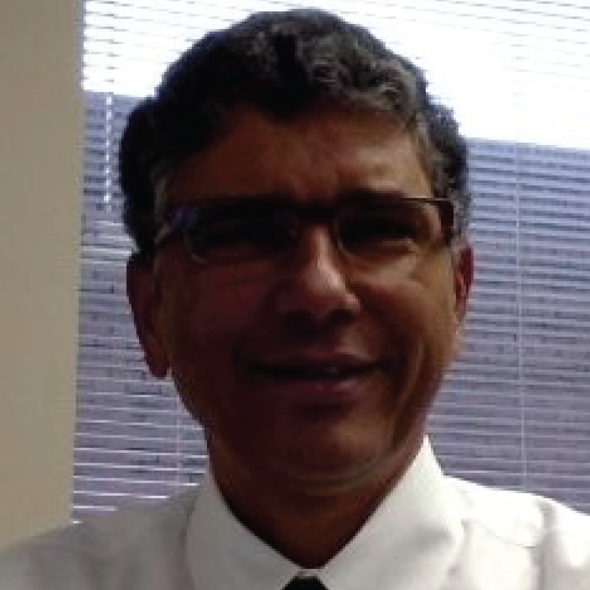 A man wearing glasses and a white shirt is smiling in front of a window.