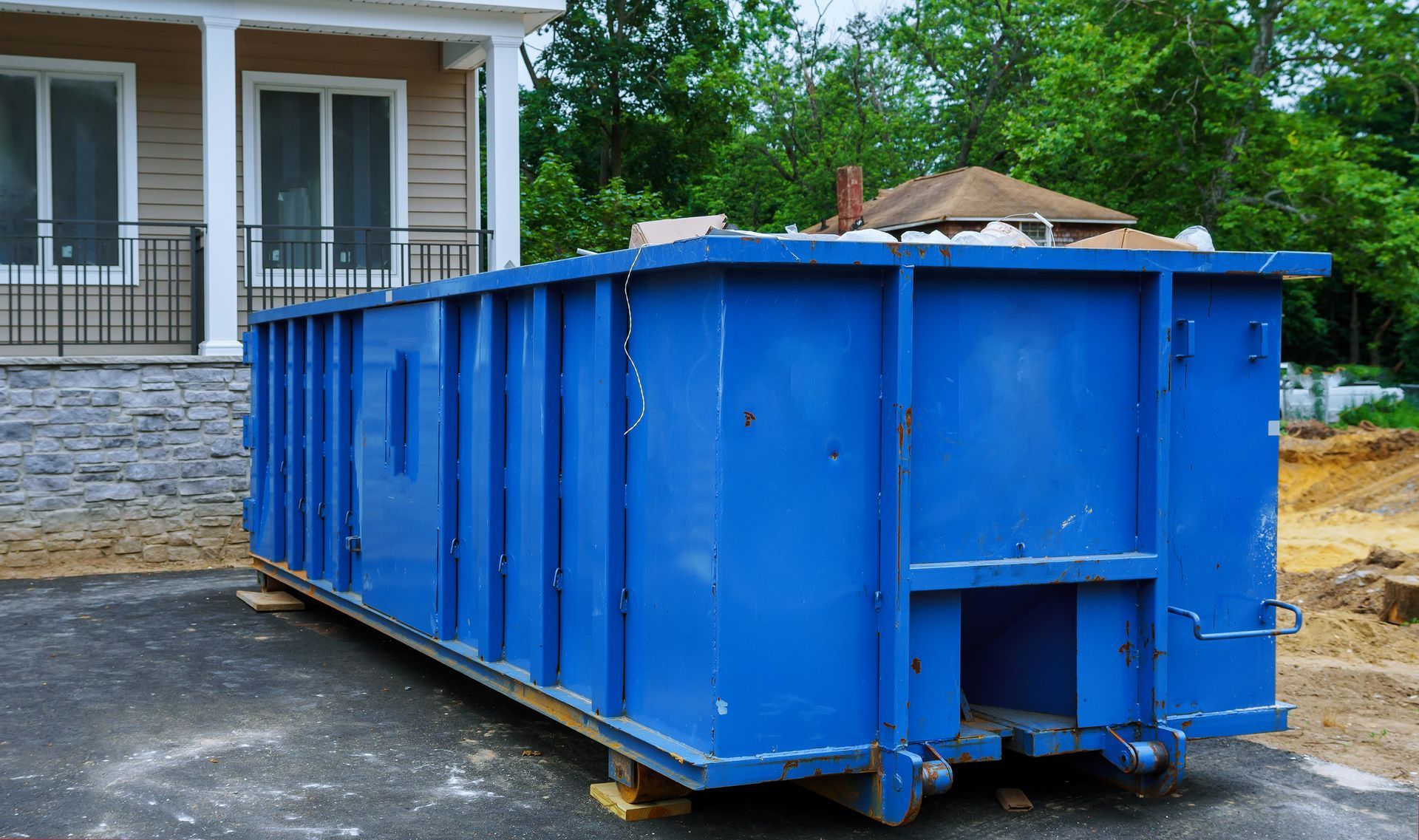 A large, bright blue dumpster sits on a paved driveway in front of a house under construction.