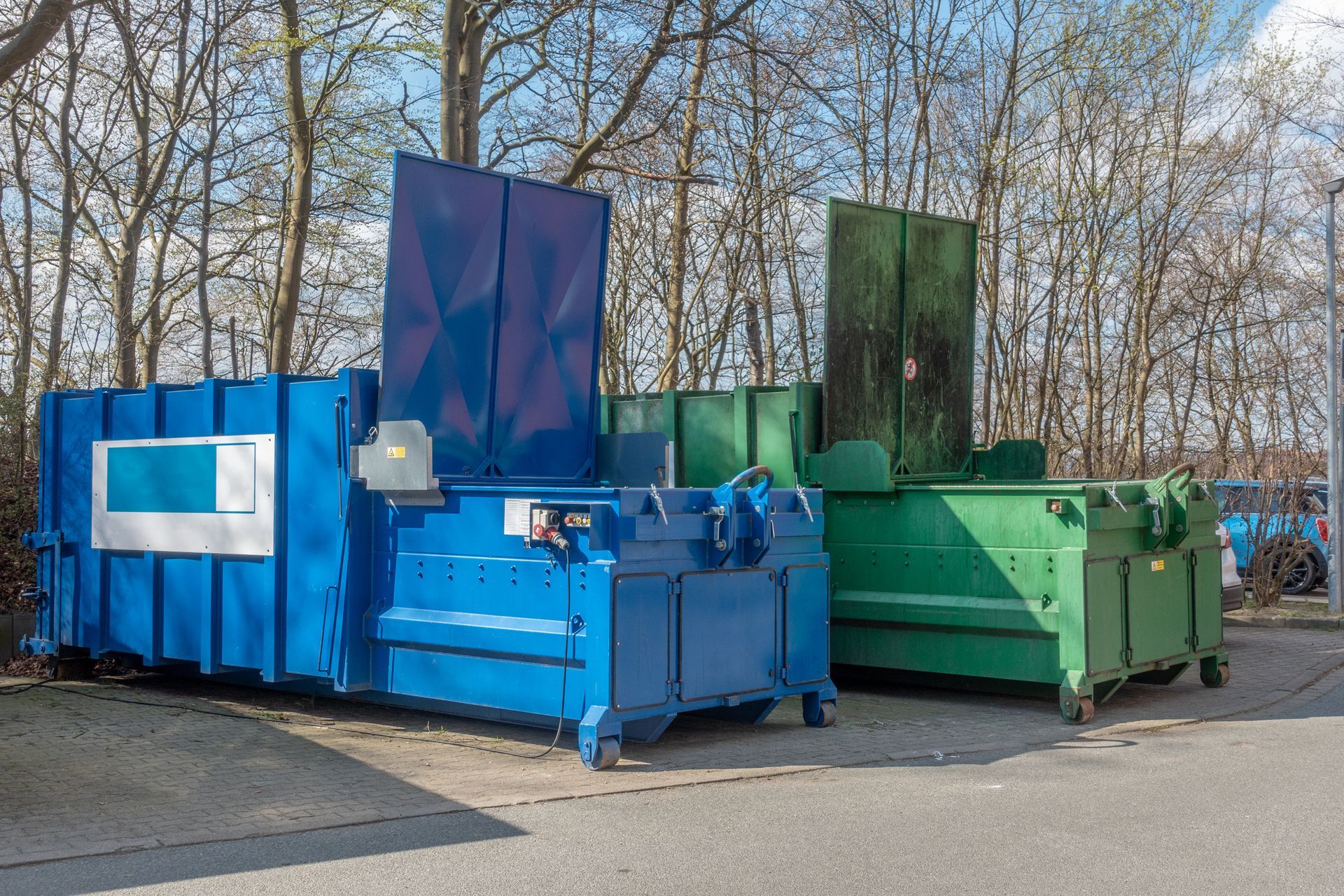 A blue and a green industrial trash compactor parked side-by-side on an asphalt lot in front of trees.