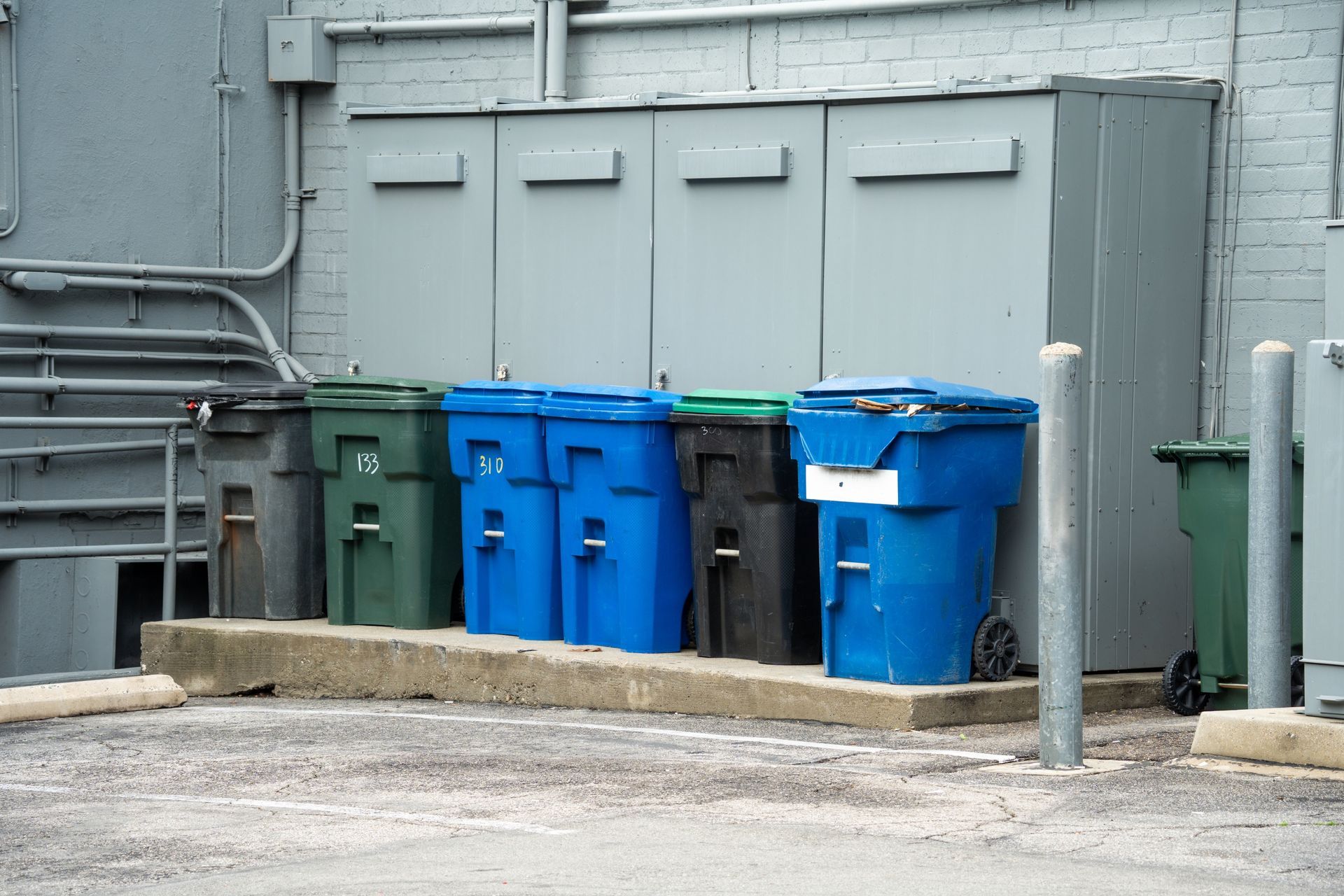 A row of green, blue, and black trash and recycling bins lined up in front of grey utility cabinets against a brick wall.