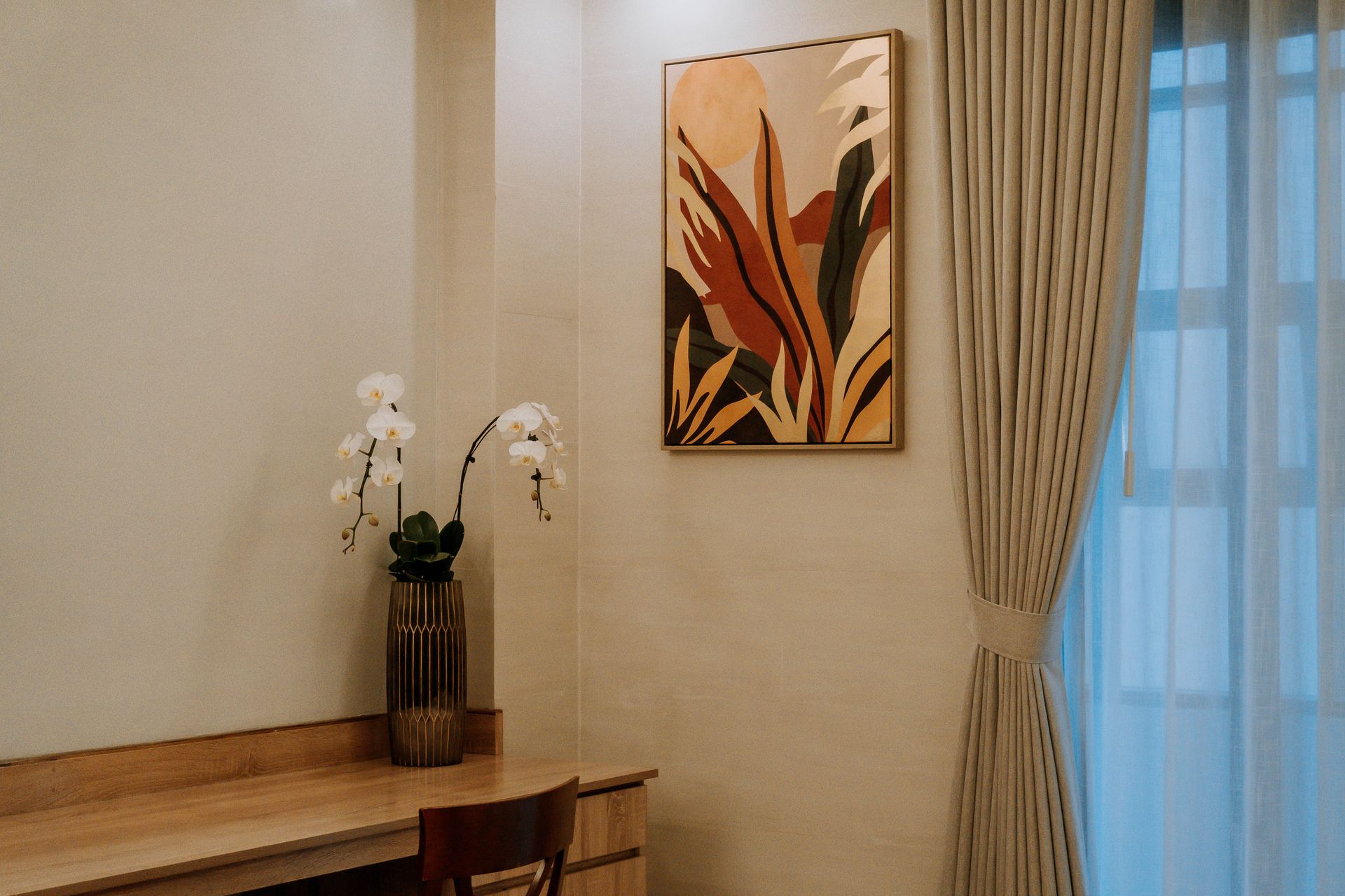 A room corner with a desk, art print of leaves, curtains, and orchids in a vase.
