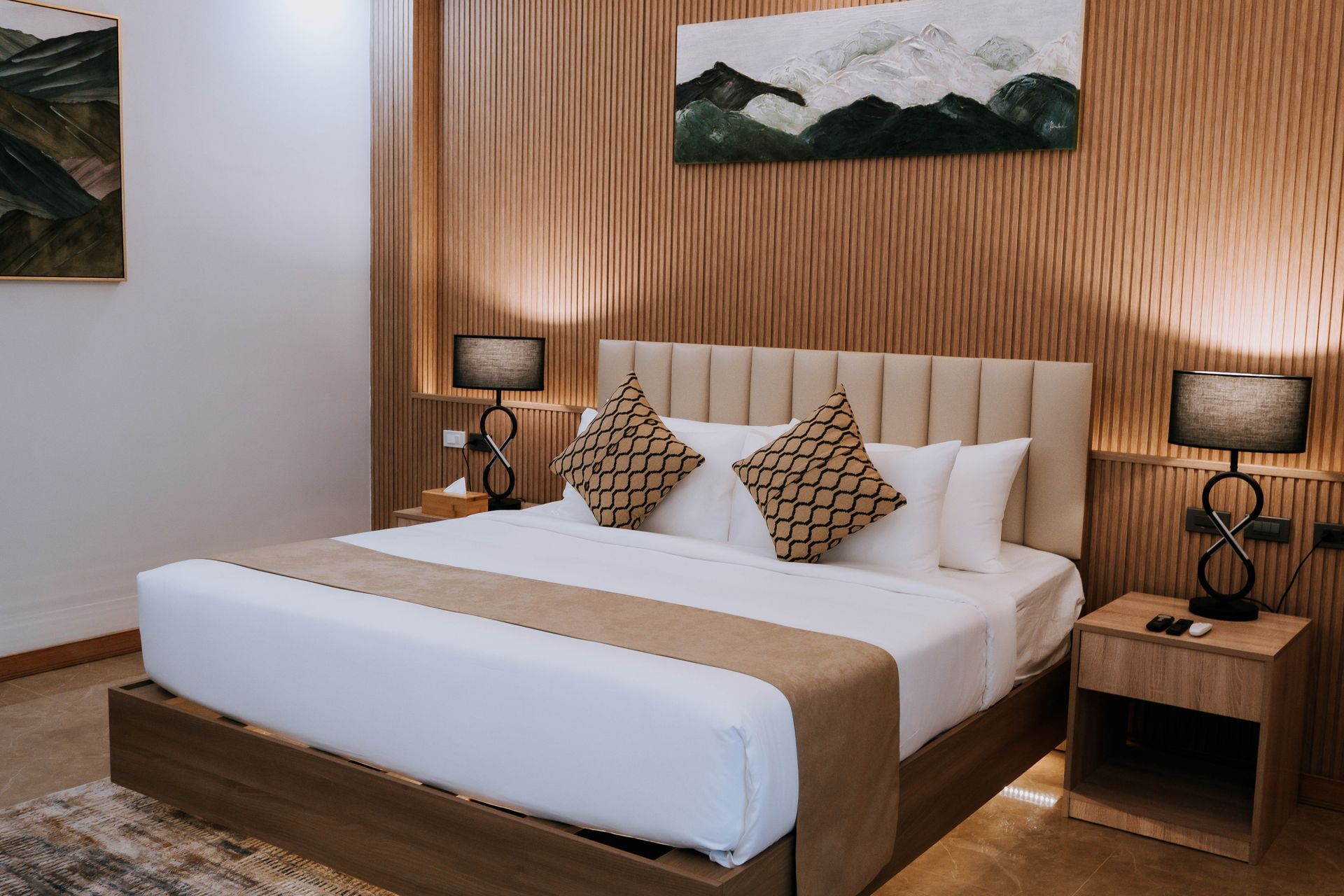 Hotel bedroom with bed, bedside tables, lamps, and art on a wood-paneled wall.