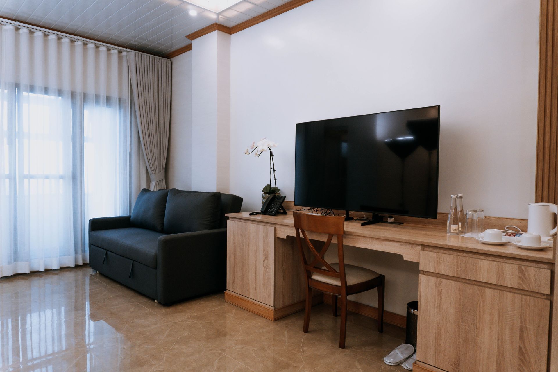 Hotel room with a black couch, desk with TV, and a window with curtains.