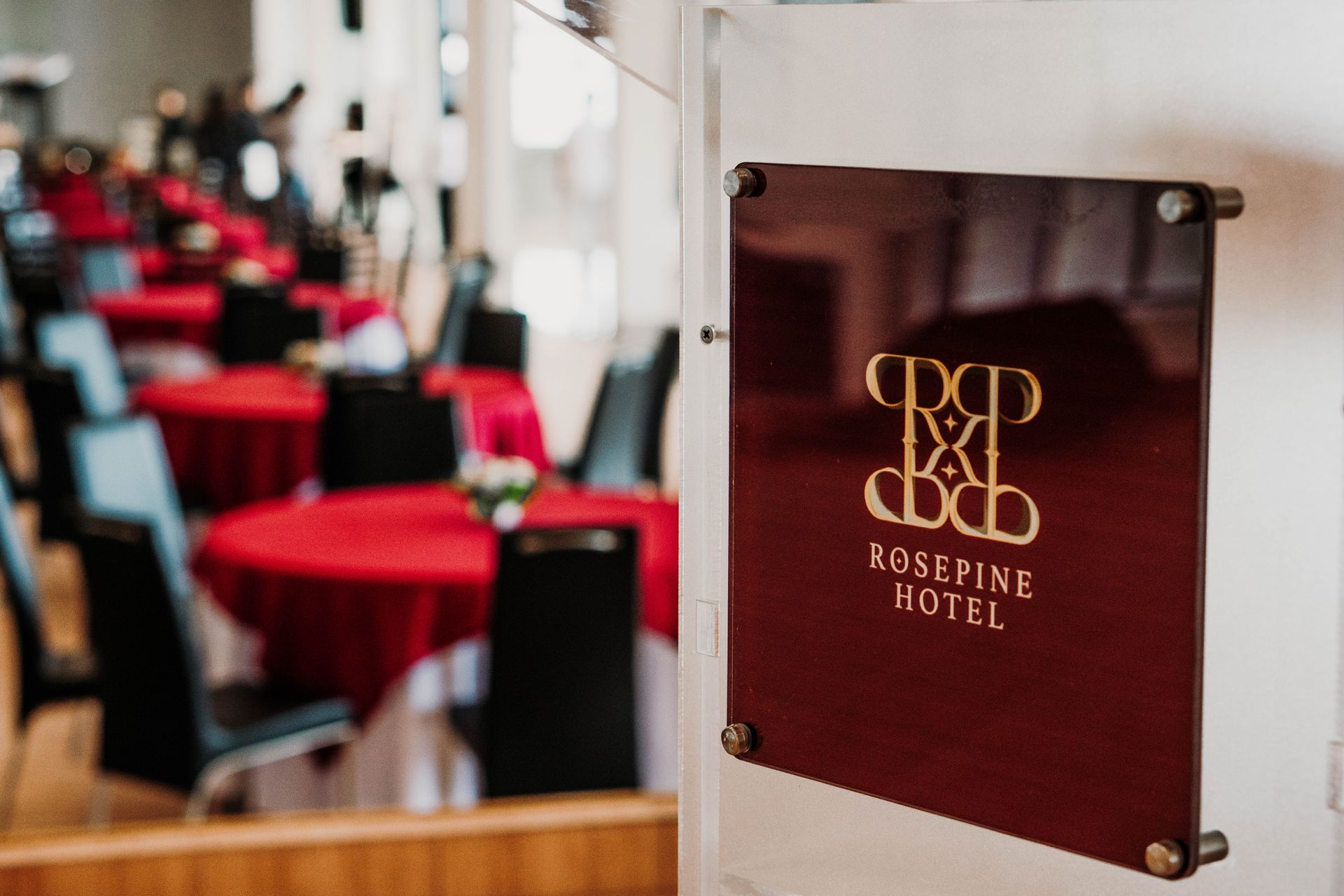 Rosepine Hotel sign, burgundy with gold logo, in front of a banquet hall with red tablecloths and black chairs.