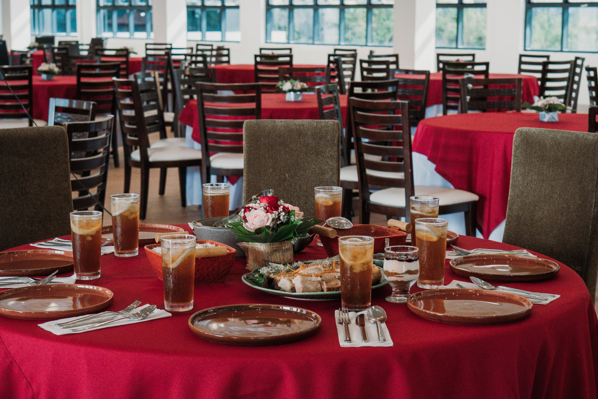 Tables set for a banquet; red tablecloths, food, drinks, and chairs arranged in a large room.